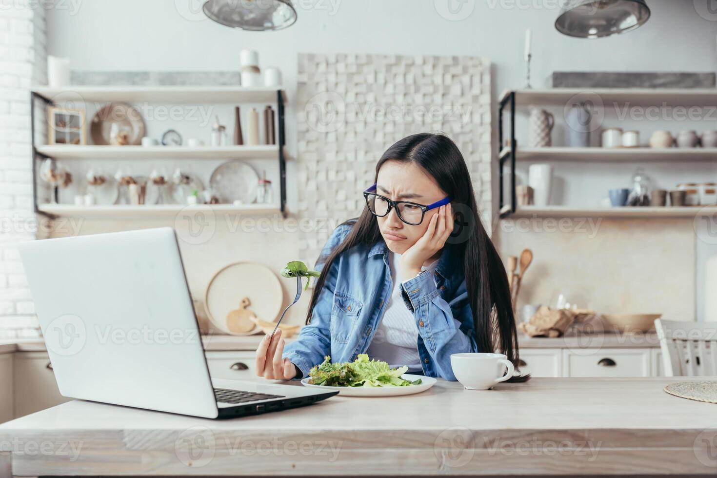 Lunch while working online. A young Asian woman sits in the kitchen at home with a laptop and unsatisfiedly eats lunch, fresh salad, vegetables. Hates diet, healthy lifestyle photo