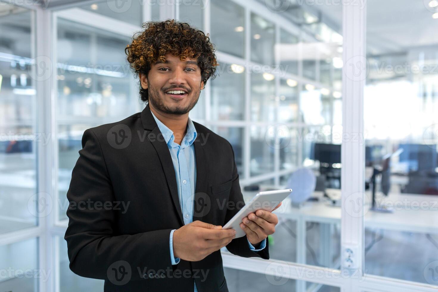 Portrait of young successful hispanic businessman, man in business suit standing inside office at workplace, businessman holding tablet computer smiling and looking at camera. photo