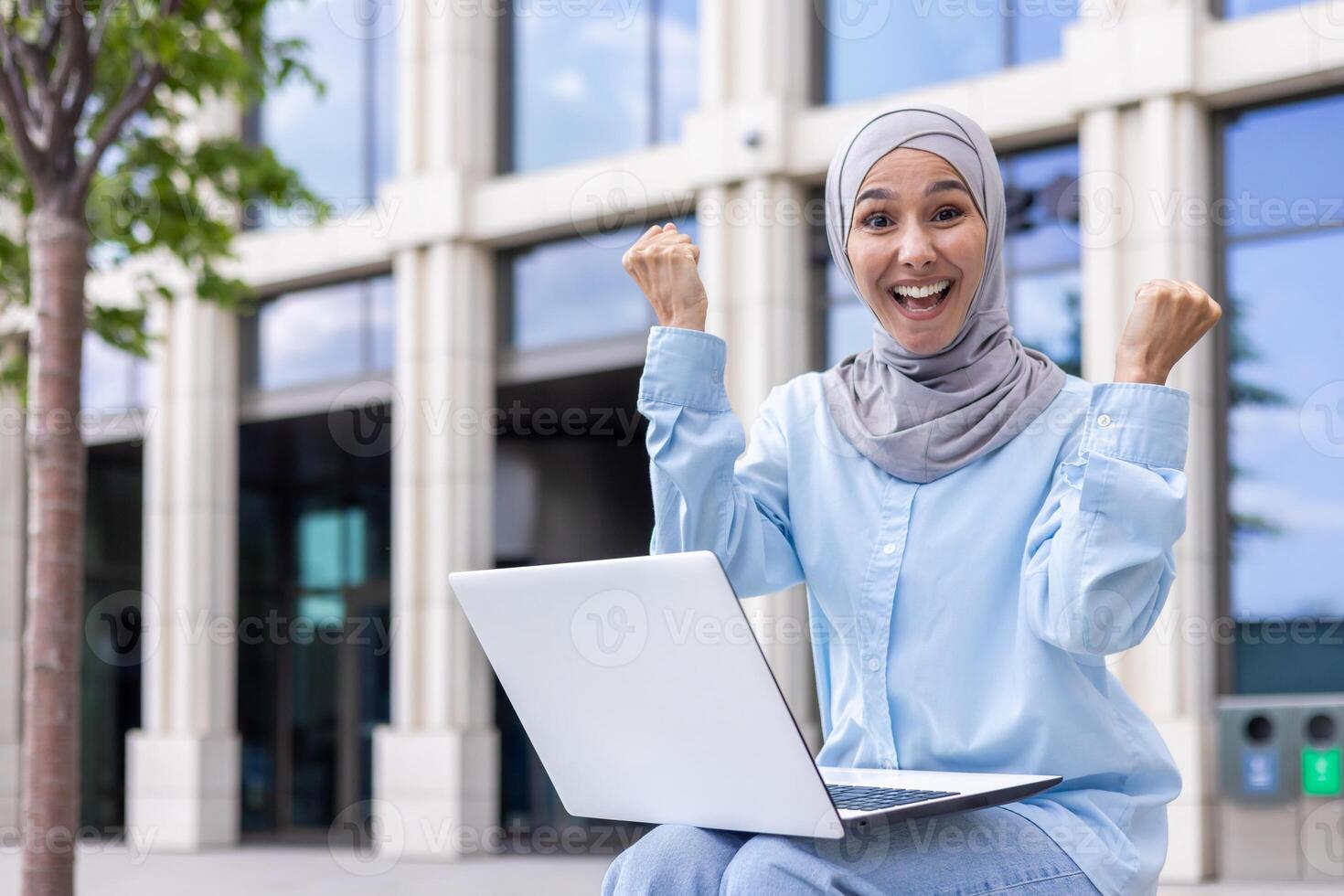 emocionado musulmán mujer en hijab se sienta al aire libre con su computadora portátil, celebrando un profesional éxito con un jubiloso expresión cerca un oficina edificio. foto