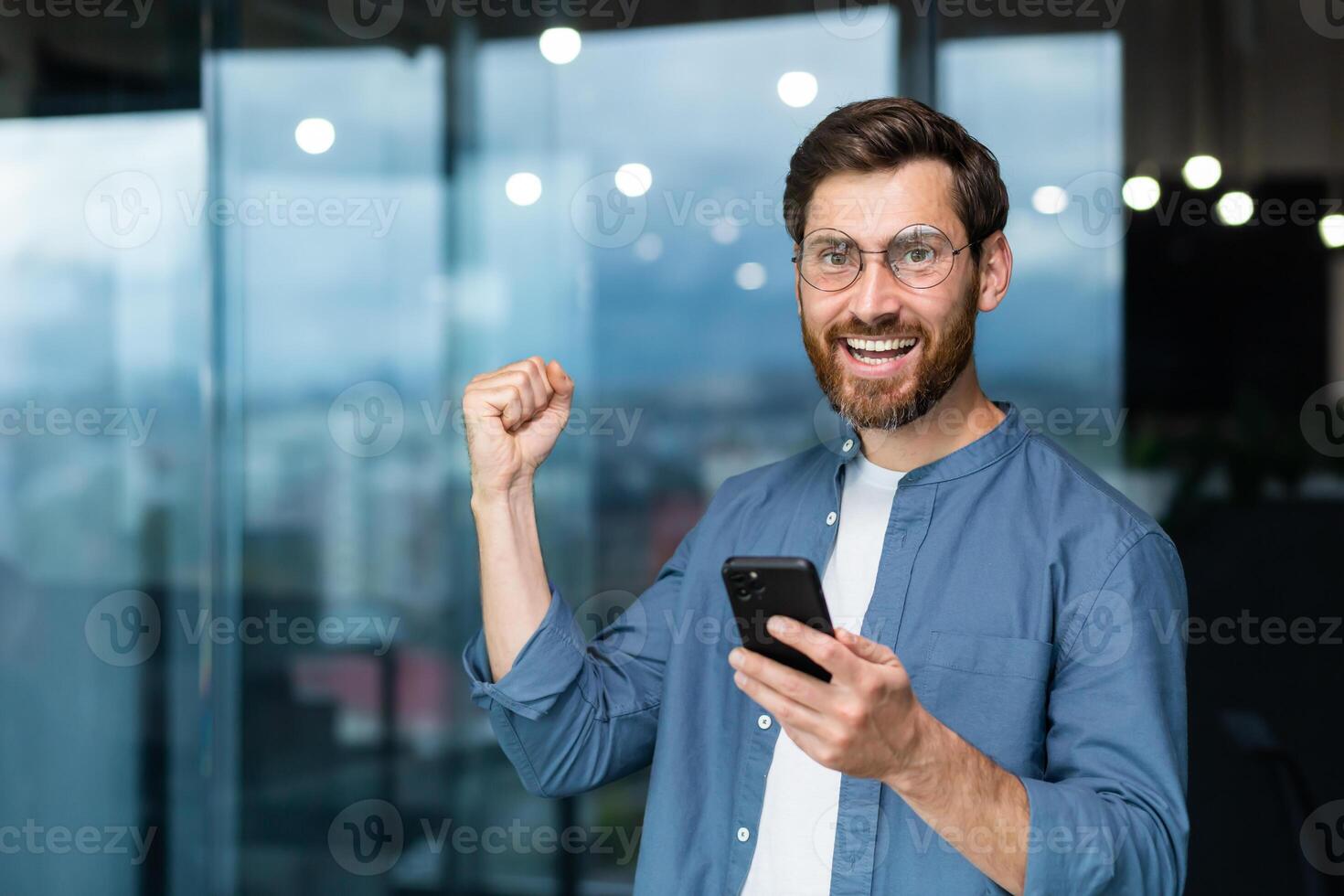 Portrait of a modern businessman in the middle of the office, the man is looking at the camera with a smartphone and celebrating the victory, the man is holding the phone in his hands and raising his hand in a gesture of triumph. photo