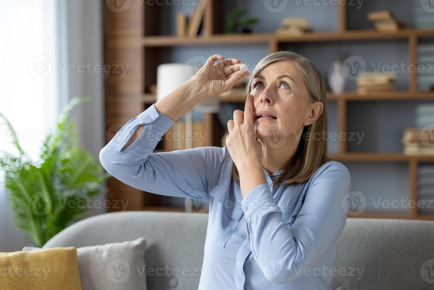 Concentrated mature lady tipping head back and fixing position for applying medical eye drops indoors. Unhealthy female with ophthalmology problems using hydrating treatment for vision care. photo
