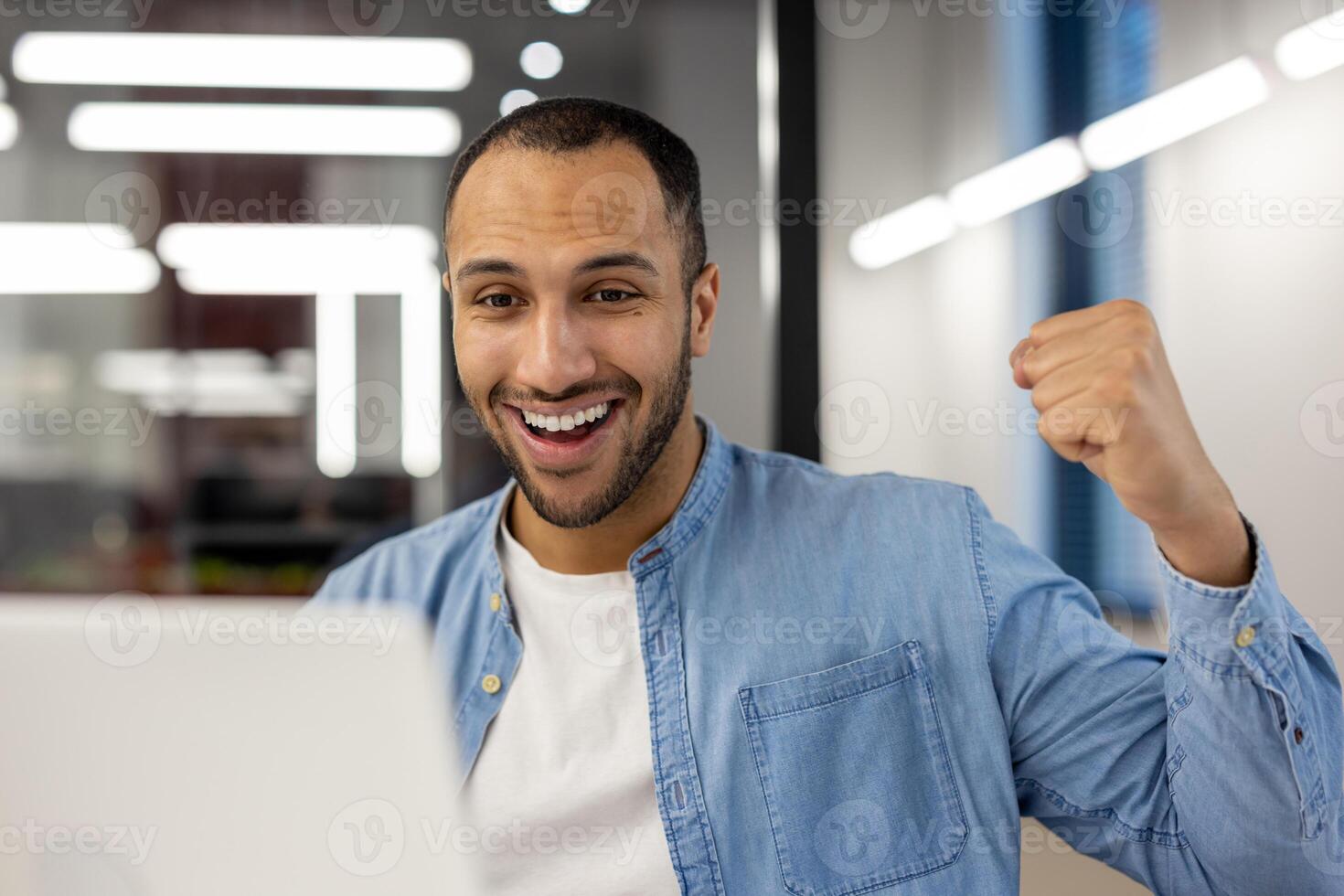 un exuberante joven hombre en un ligero azul mezclilla camisa puñetazos el aire con alegría en frente de su ordenador portátil a el oficina, simbolizando profesional éxito y logro. foto