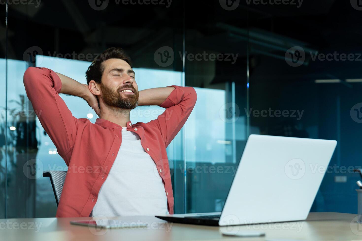 exitoso empresario en oficina en un rojo camisa descansa con su manos doblada cabeza con cerrado ojos, maduro hombre con un barba trabajos con un ordenador portátil a el lugar de trabajo adentro, Sueños de futuro logros foto