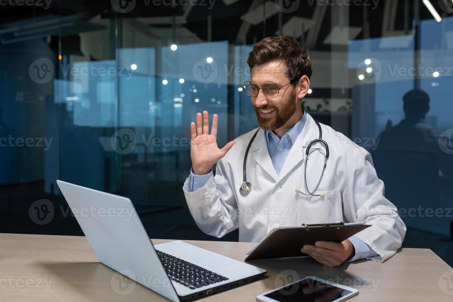 Mature doctor working remotely from modern clinic office, man inside talking on call with patient using laptop photo