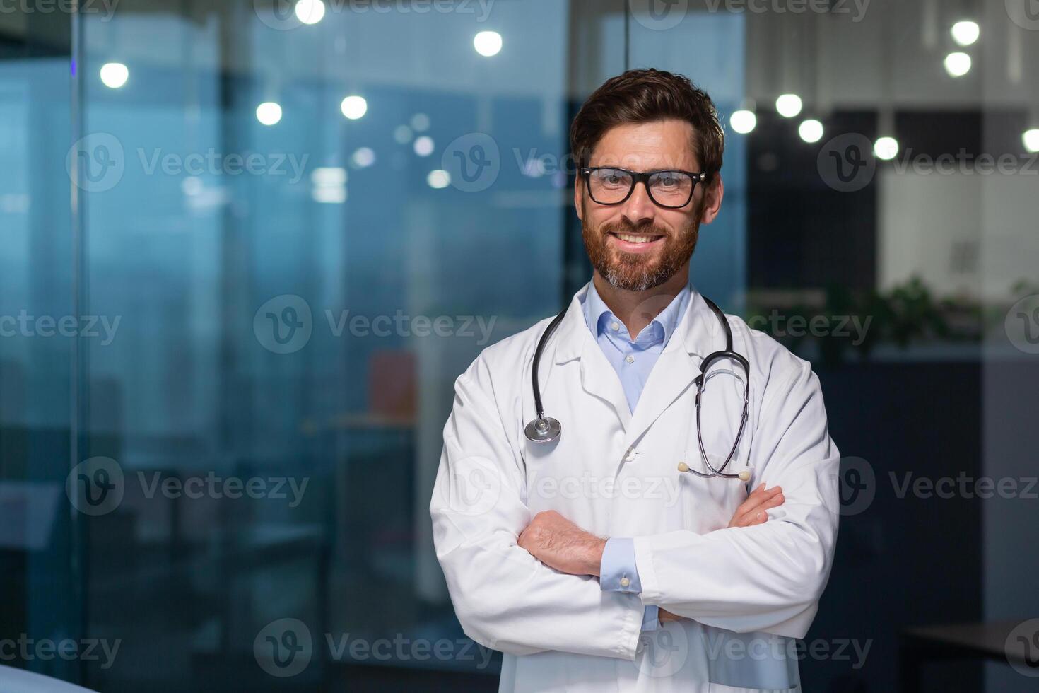 Portrait of mature doctor with beard, man in white medical coat smiling and looking at camera with crossed arms working inside modern clinic. photo