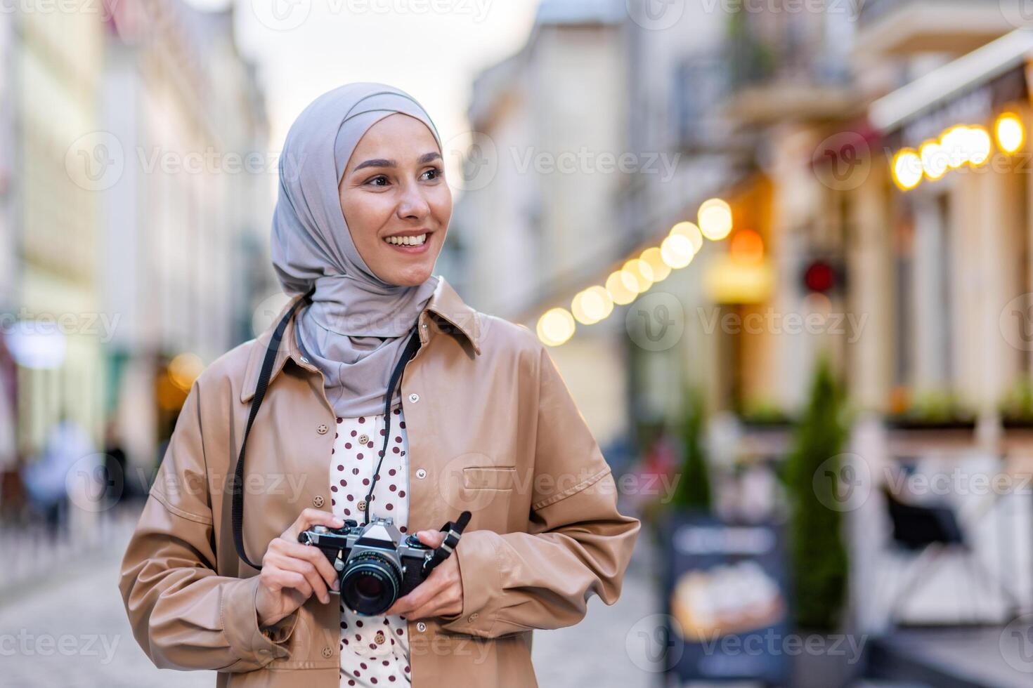 Young beautiful woman walking in the evening city in hijab, tourist with camera and wearing a hat inspects the historical city smiling with satisfaction, Muslim woman on a trip. photo