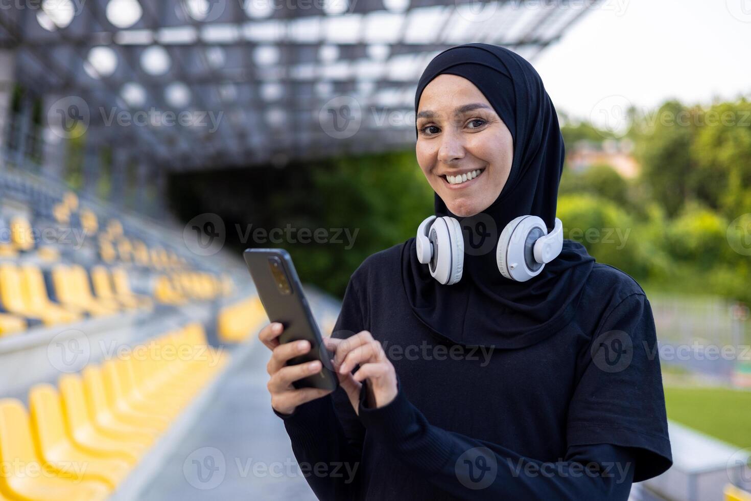 Portrait of a young hijab Muslim woman in a sports stadium running and doing active physical exercises, smiling and looking at the camera, using the phone and headphones to listen to music. photo