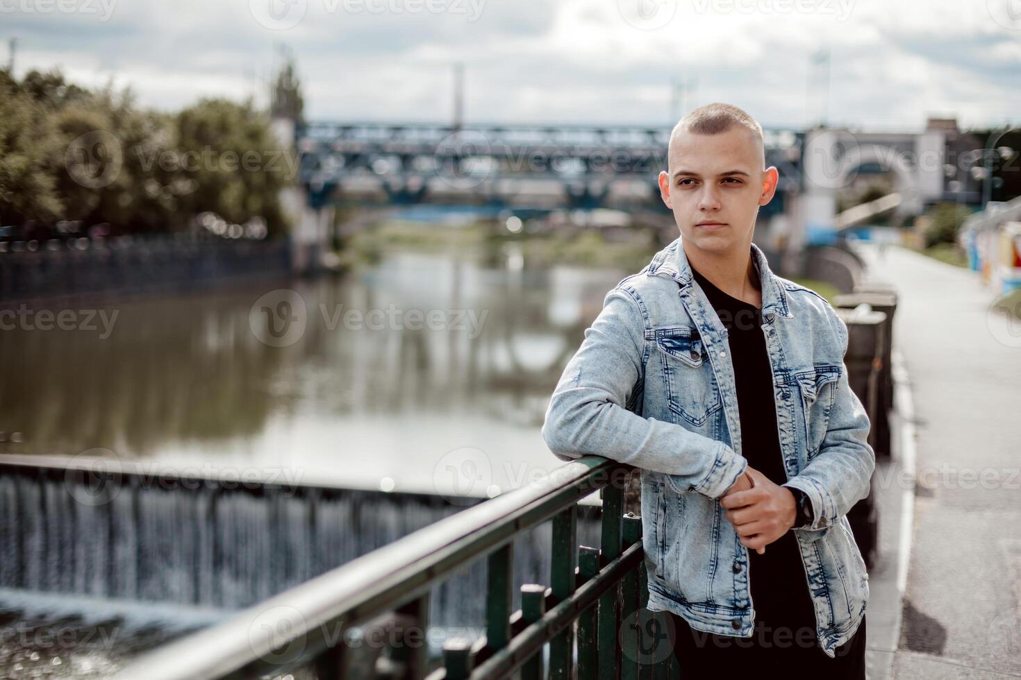 A young man stands on a bridge, wearing a blue jacket and a black shirt photo