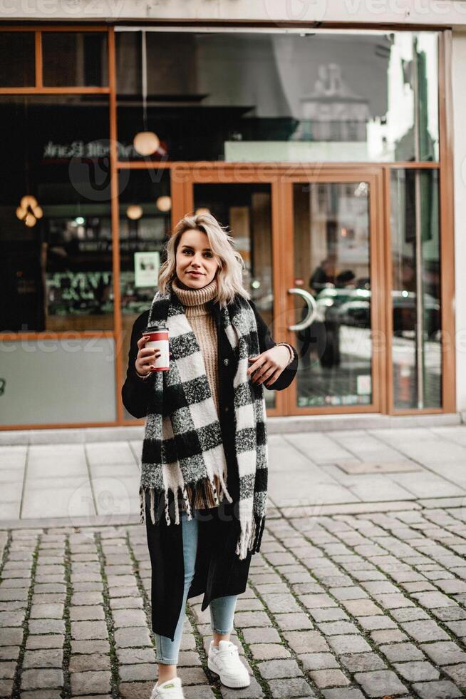 A stylish woman stands in front of a building, clutching a vibrant red coffee cup. Her black coat and plaid scarf add flair to the urban scene, capturing the essence of a brisk city morning photo