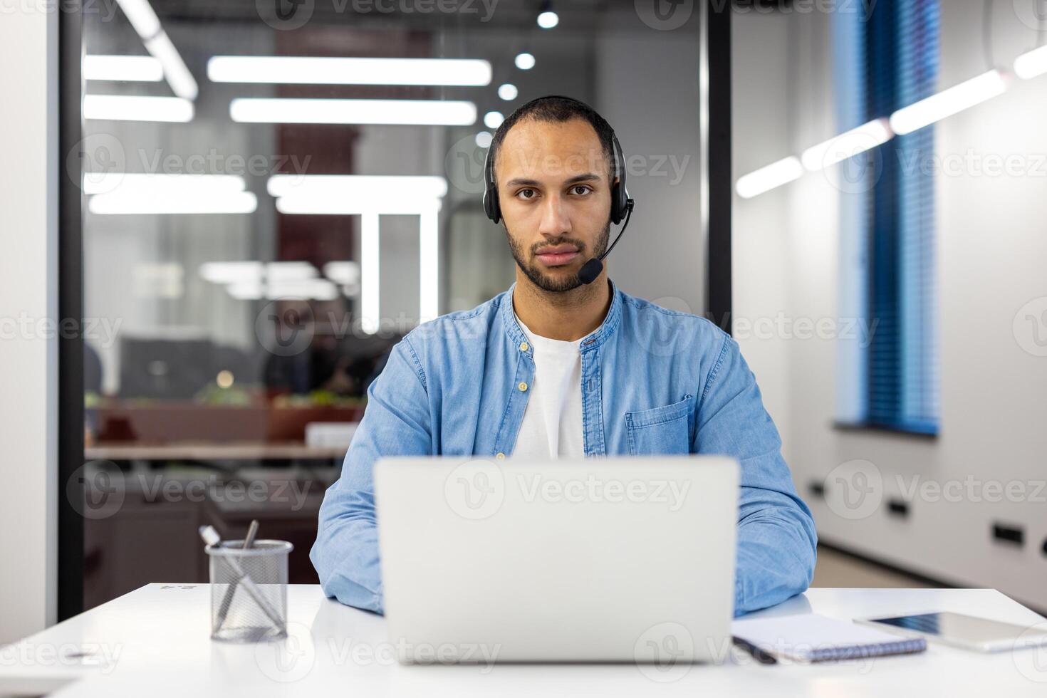 Portrait of a serious young hispanic male specialist wearing a headset, sitting at a desk in a busy office, working on a laptop, looking intently at the camera. photo