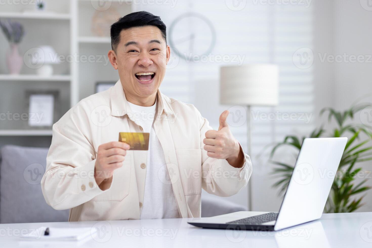 Cheerful asian man making like sign with hand while sitting at domestic office with laptop and plastic bank card. Emotional remote programmer receiving financial bonus for productive monthly work. photo