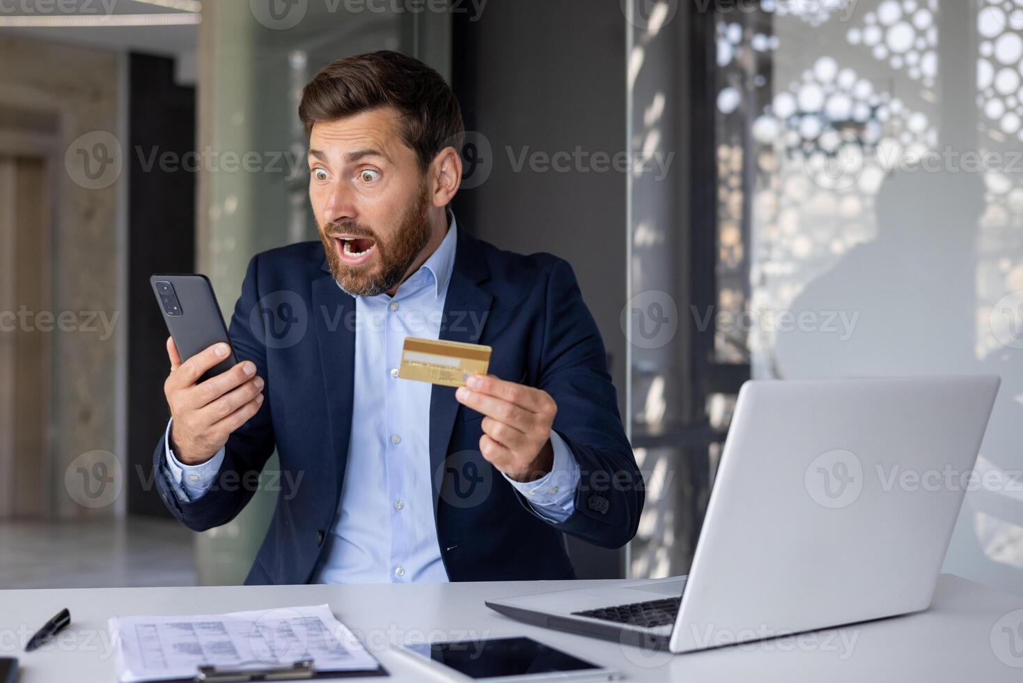 Shocked young man in business suit sitting at desk in office, holding credit card in hand and looking surprised at mobile phone screen. photo