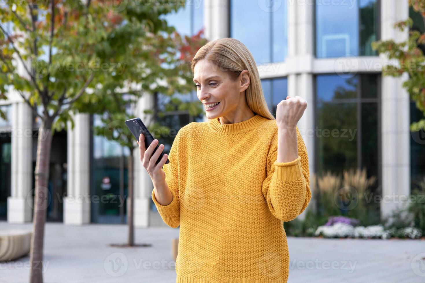 A happy middle-aged woman in a yellow sweater celebrates while looking at her smartphone outside, with modern buildings and autumn trees in the background. photo