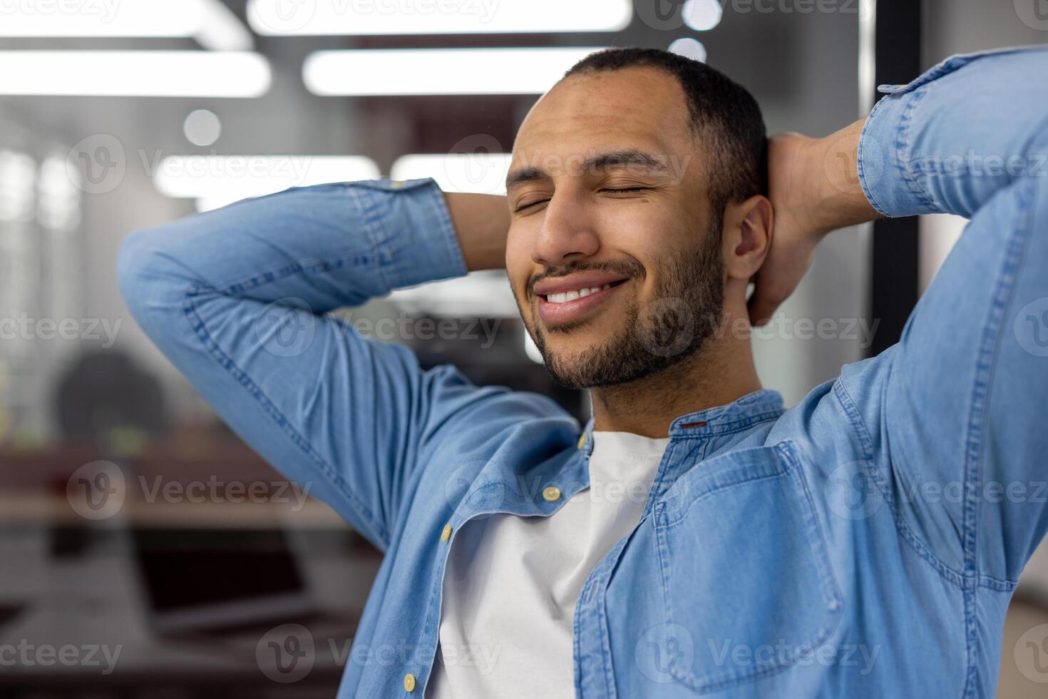 A close-up photo of a young Latin American man sitting contentedly and smiling in a modern office with his eyes closed and his arms, relaxed behind his head.