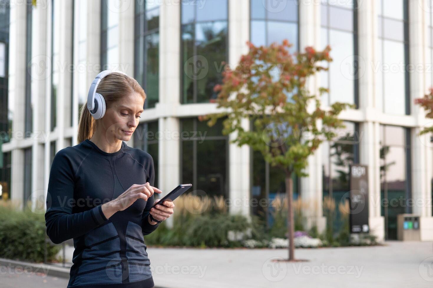 un enfocado mujer disfrutando su lista de reproducción mientras manejo su teléfono inteligente en un sin prisa postura, vestido en aptitud atuendo, con moderno arquitectura y verdor en el antecedentes. foto