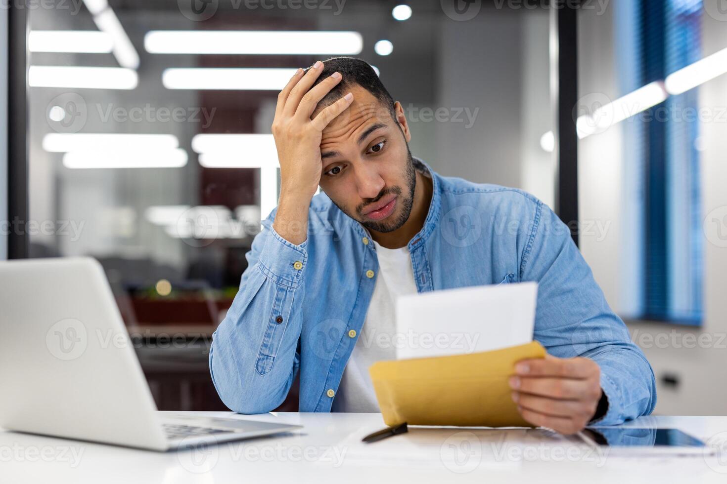 Concerned Indian man in casual attire sits at home, analyzing a letter. Expression of stress and anxiety while reading an envelope's contents in a living room. photo