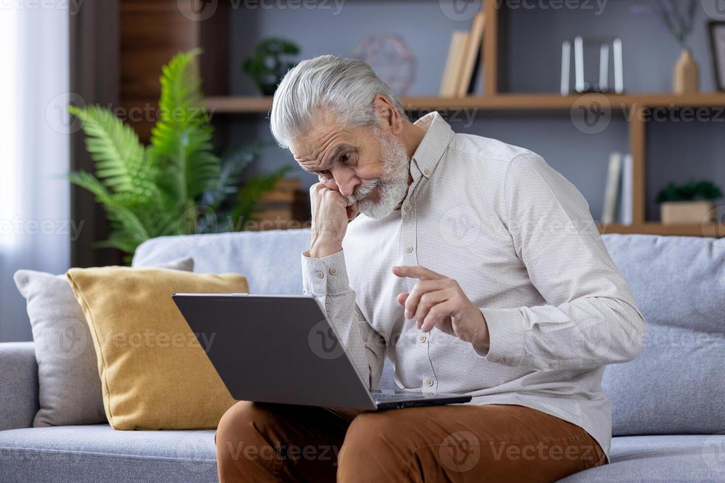 enfocado mayor hombre con blanco pelo y barba profundamente absorto en utilizando un ordenador portátil mientras sentado en un sofá en un bien iluminado vivo habitación, representando moderno tecnología utilizar por personas mayores foto