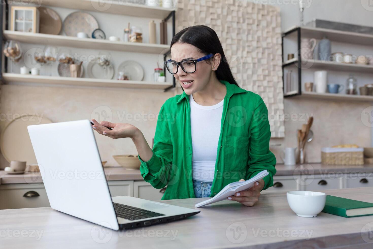Angry young woman working from home. Talking on a call from a laptop, writes in a notebook photo