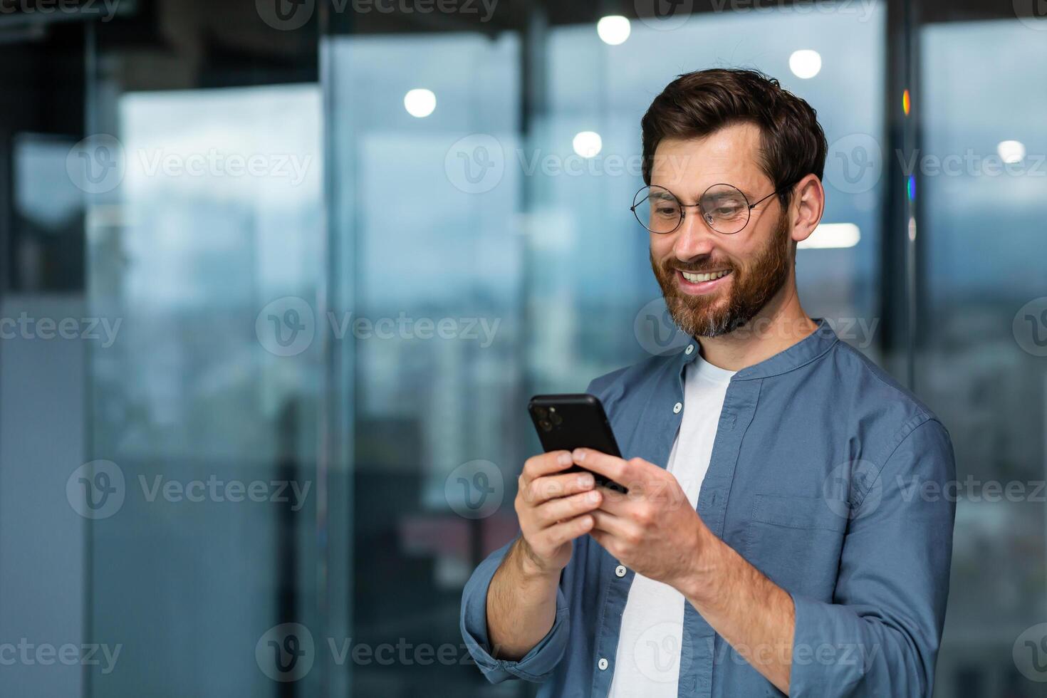 Successful businessman in glasses and beard standing near window in the evening, man using phone, happy boss typing message and browsing online pages on smartphone app inside office. photo
