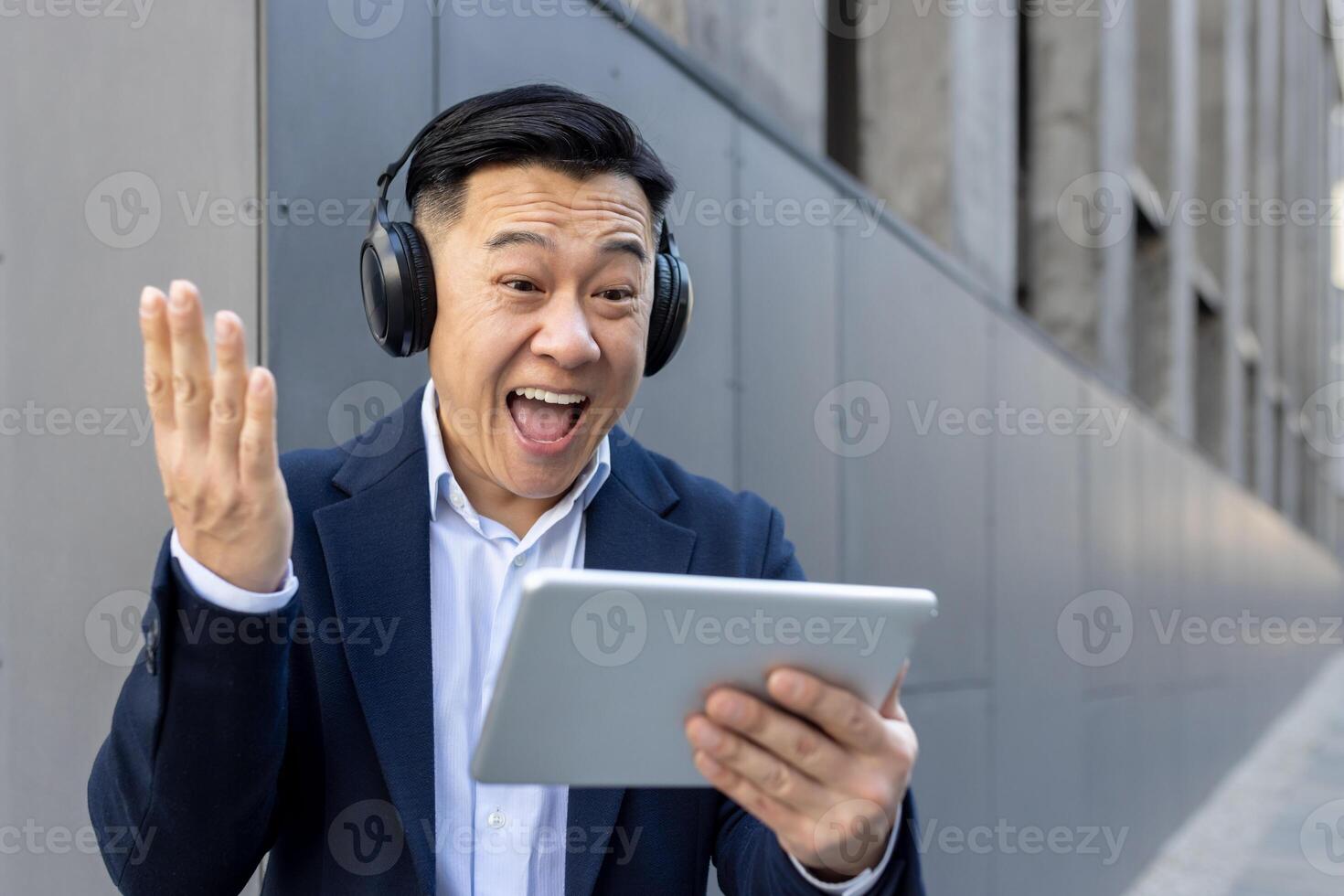 contento joven asiático masculino empresario en pie en auriculares fuera de oficina centro, mirando a tableta pantalla y contento con Noticias y éxito. foto