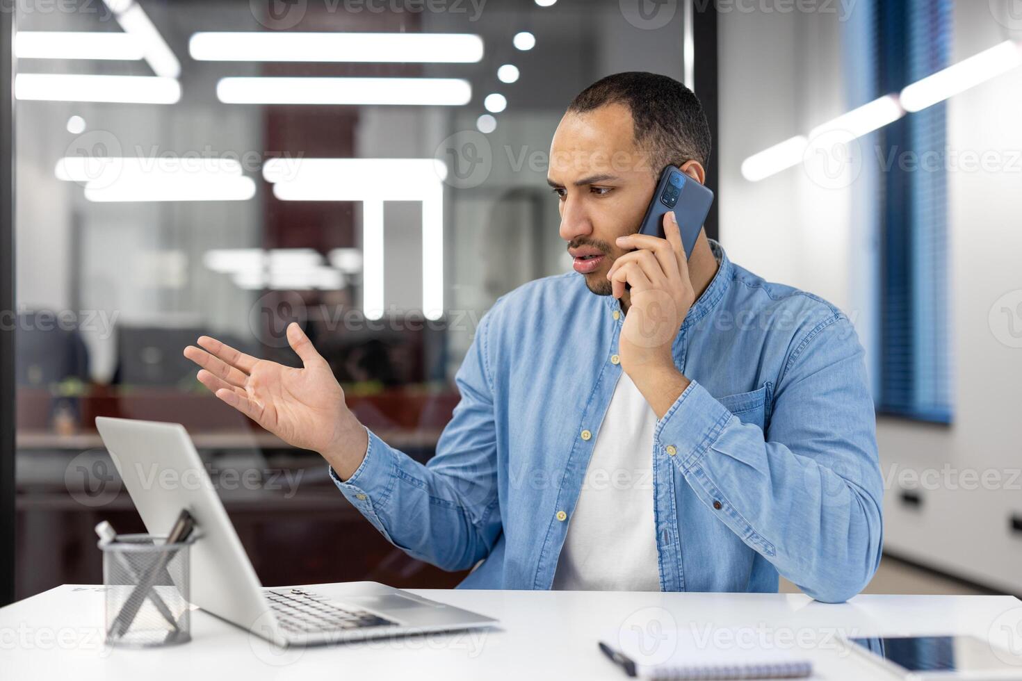 A man is talking on his cell phone while sitting at a desk. He is wearing a blue shirt and white shirt. There is a laptop on the desk in front of him photo