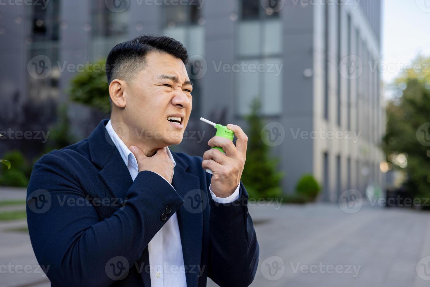 Asian young businessman sitting near working office outside, holding throat with hand, feeling severe pain and using medical spray. photo