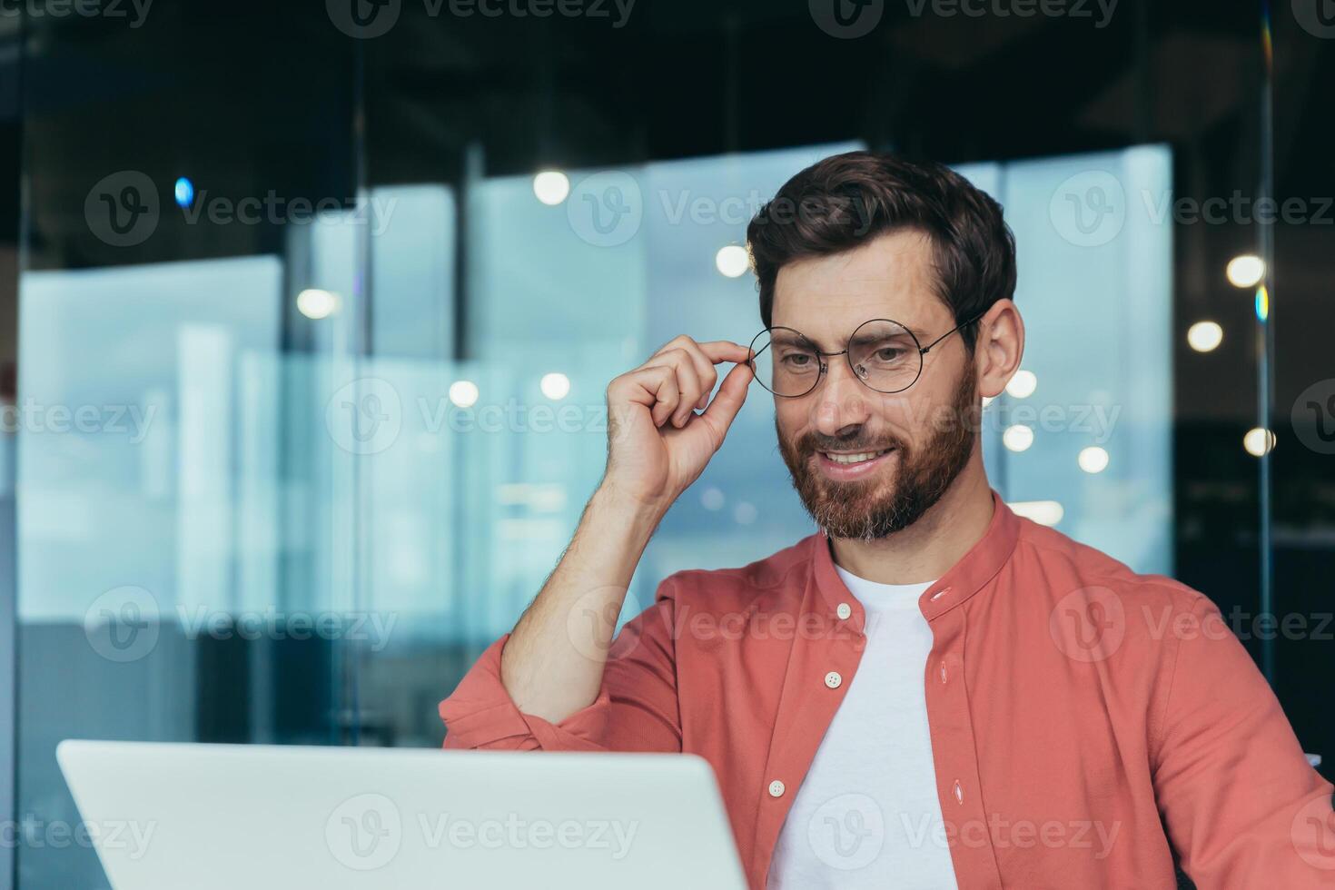Close-up photo of successful and happy architect working inside modern design studio studio, man in glasses and red shirt smiling and looking at laptop screen.
