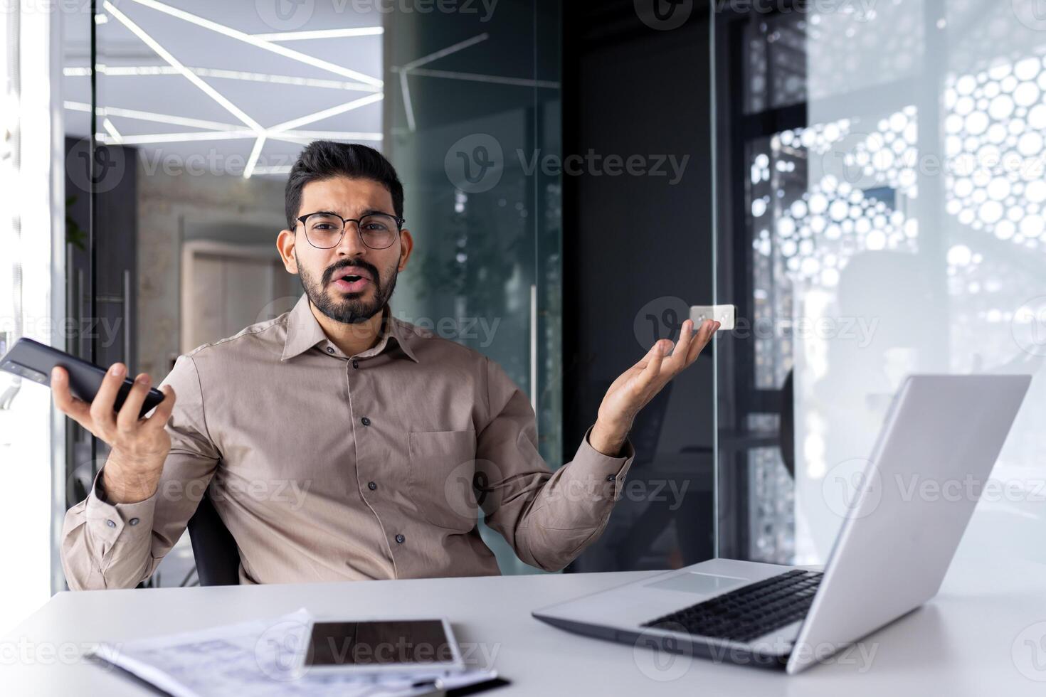 Portrait of a young angry and disappointed Indian man sitting at an office desk, holding a mobile phone and spreading his hands to the camera. photo