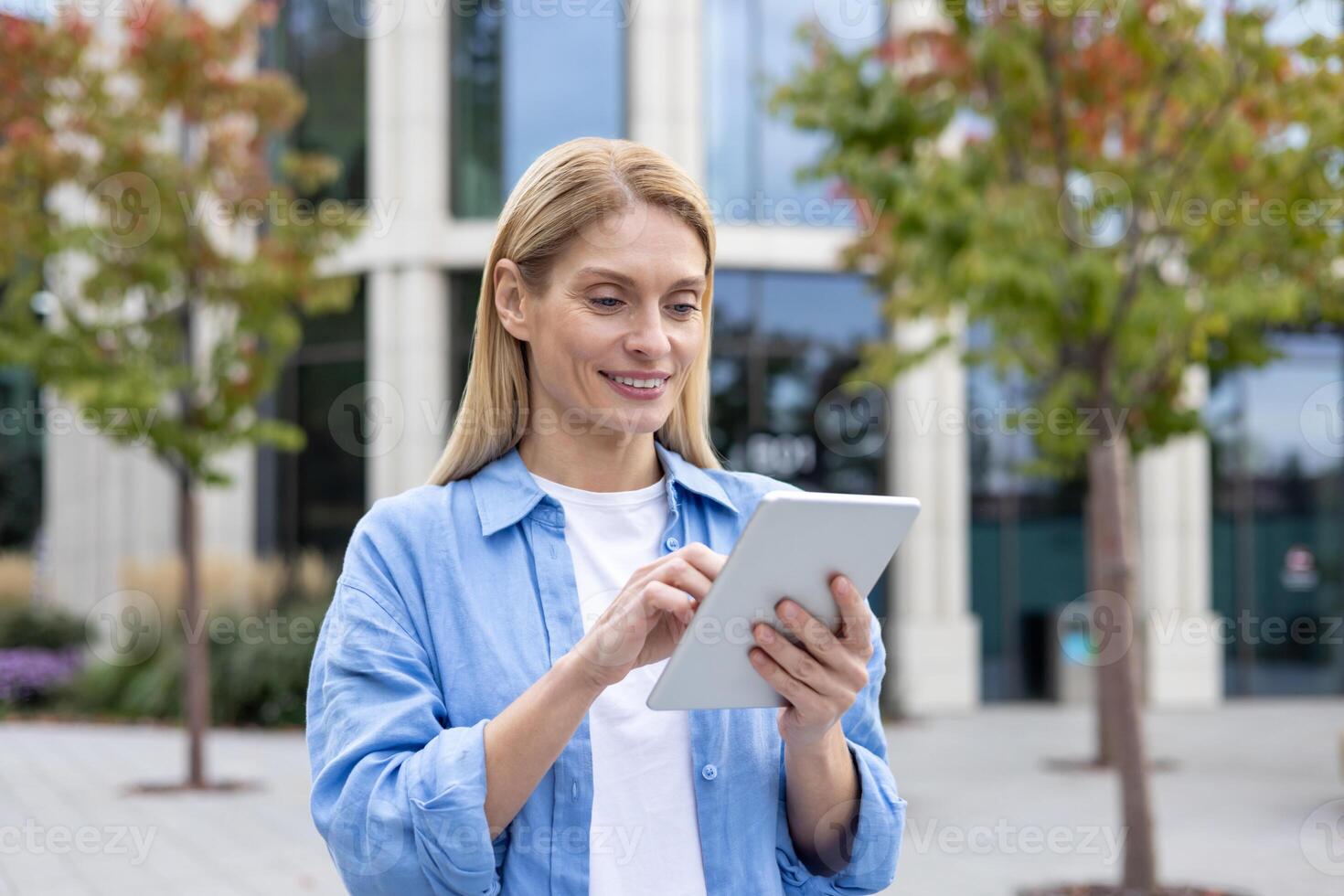 Smiling blonde woman using digital tablet while standing outdoors at pedestrian space with trees. Confident landscape designer developing green zone area in district with futuristic skyscrapers. photo