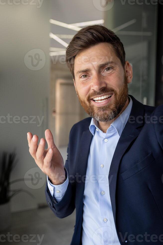 Vertical close-up photo of a young businessman in a suit who is in the office, holding a camera and talking on a call, smiling and gesturing with his hands.