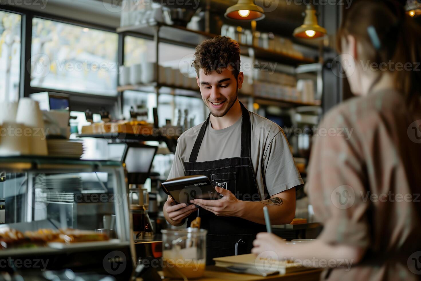 young man in a coffee shop stands behind the cashier counter speaking with a customer holding a tablet photo