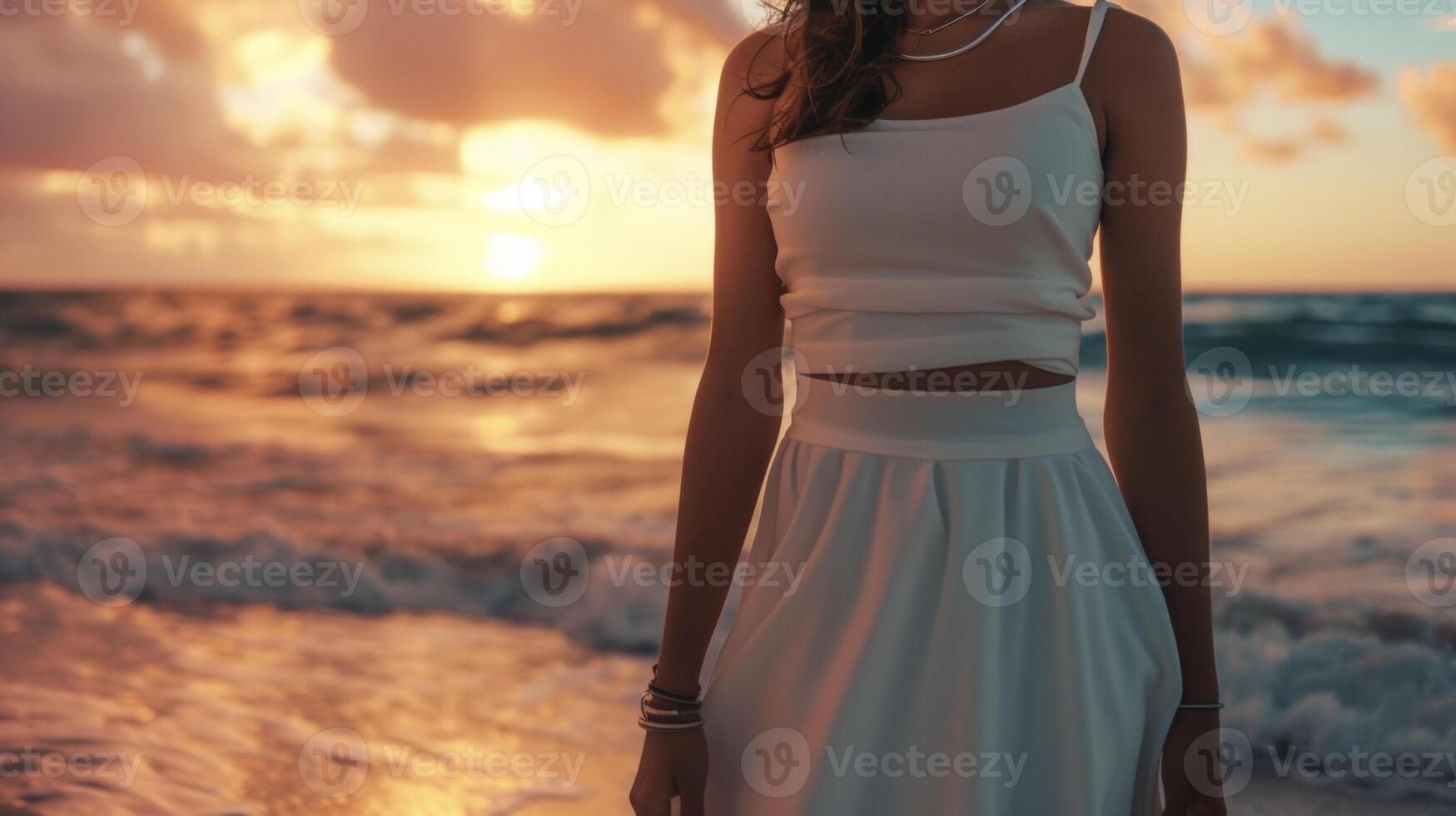 A sleek white midi dress in a figureflattering is accented with minimalistic silver jewelry. The background is a beach at sunset with the sky turning shades of pink and orange photo