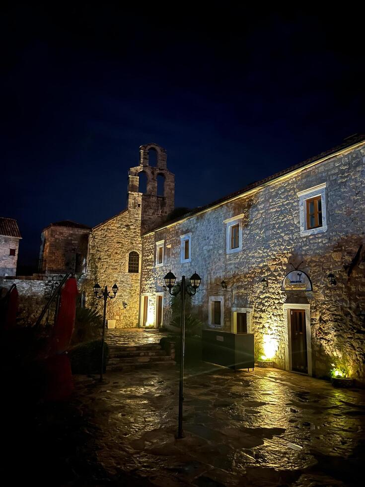 buda, montenegro - 25 diciembre 2022. patio de un antiguo Roca Iglesia con un campana torre a noche en el iluminación de linternas foto