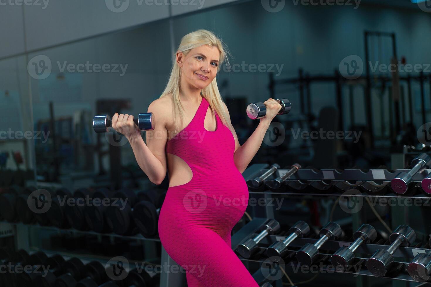 embarazada mujer levantamiento arriba pesas formación bíceps músculo a el gimnasio en pie cerca espejo foto
