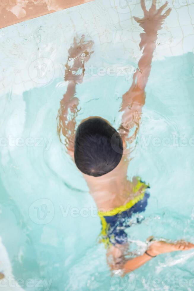Happy Indian boy swimming in a pool, Kid wearing swimming costume along with air tube during hot summer vacations, Children boy in big swimming pool. photo
