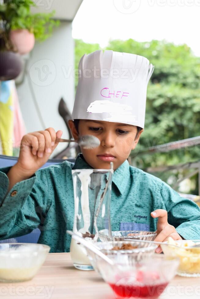 Cute Indian chef boy preparing sundae dish as a part of non fire cooking which includes vanilla ice cream, brownie, coco powder, freshly chopped fruits and strawberry syrup. Little kid preparing food photo