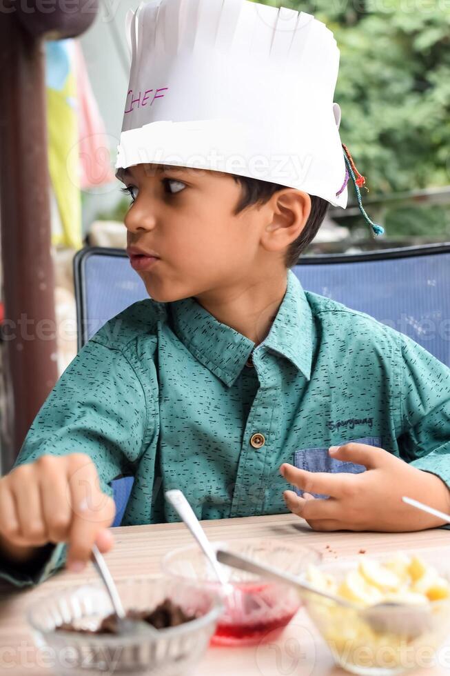 Cute Indian chef boy preparing sundae dish as a part of non fire cooking which includes vanilla ice cream, brownie, coco powder, freshly chopped fruits and strawberry syrup. Little kid preparing food photo