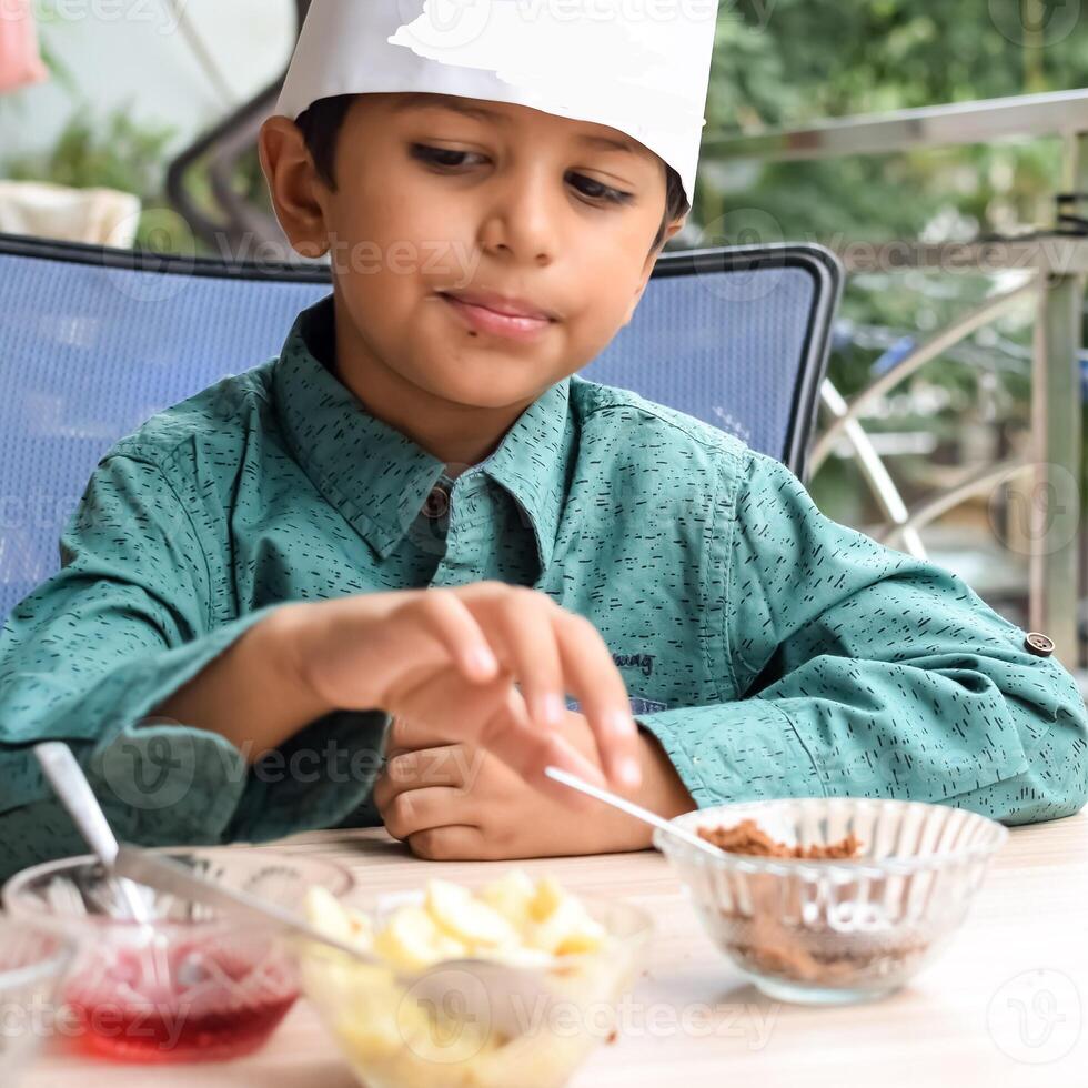 Cute Indian chef boy preparing sundae dish as a part of non fire cooking which includes vanilla ice cream, brownie, coco powder, freshly chopped fruits and strawberry syrup. Little kid preparing food photo