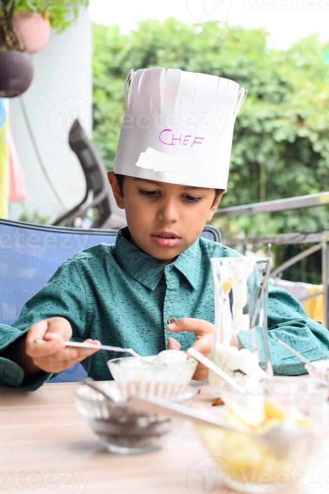 Cute Indian chef boy preparing sundae dish as a part of non fire cooking which includes vanilla ice cream, brownie, coco powder, freshly chopped fruits and strawberry syrup. Little kid preparing food photo