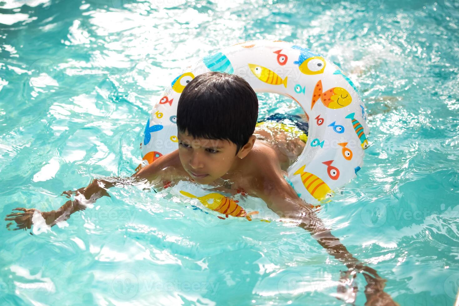 Happy Indian boy swimming in a pool, Kid wearing swimming costume along with air tube during hot summer vacations, Children boy in big swimming pool. photo