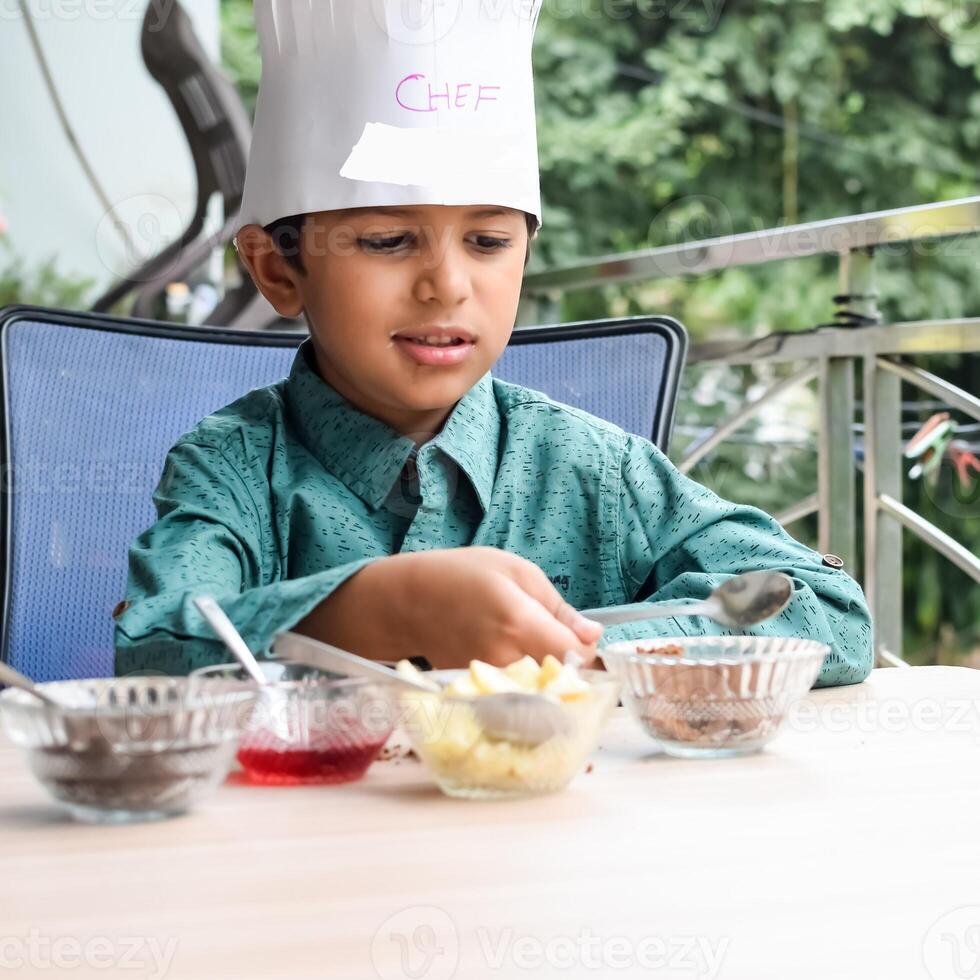 Cute Indian chef boy preparing sundae dish as a part of non fire cooking which includes vanilla ice cream, brownie, coco powder, freshly chopped fruits and strawberry syrup. Little kid preparing food photo