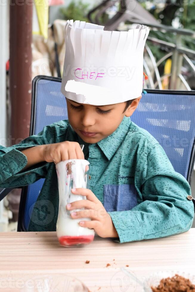 Cute Indian chef boy preparing sundae dish as a part of non fire cooking which includes vanilla ice cream, brownie, coco powder, freshly chopped fruits and strawberry syrup. Little kid preparing food photo