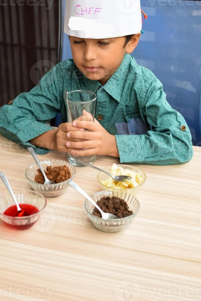 Cute Indian chef boy preparing sundae dish as a part of non fire cooking which includes vanilla ice cream, brownie, coco powder, freshly chopped fruits and strawberry syrup. Little kid preparing food photo