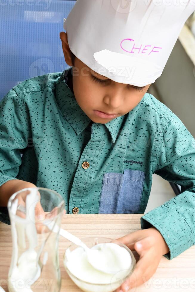 Cute Indian chef boy preparing sundae dish as a part of non fire cooking which includes vanilla ice cream, brownie, coco powder, freshly chopped fruits and strawberry syrup. Little kid preparing food photo