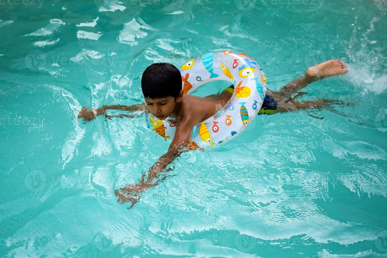 Happy Indian boy swimming in a pool, Kid wearing swimming costume along with air tube during hot summer vacations, Children boy in big swimming pool. photo