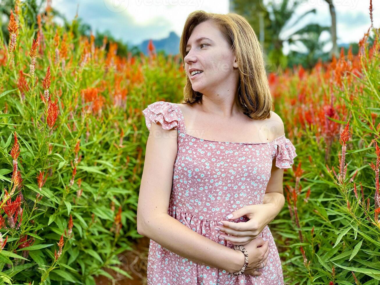 Smiling young woman in a floral summer dress standing in a vibrant red flower field with a scenic mountain backdrop, capturing the essence of summer photo