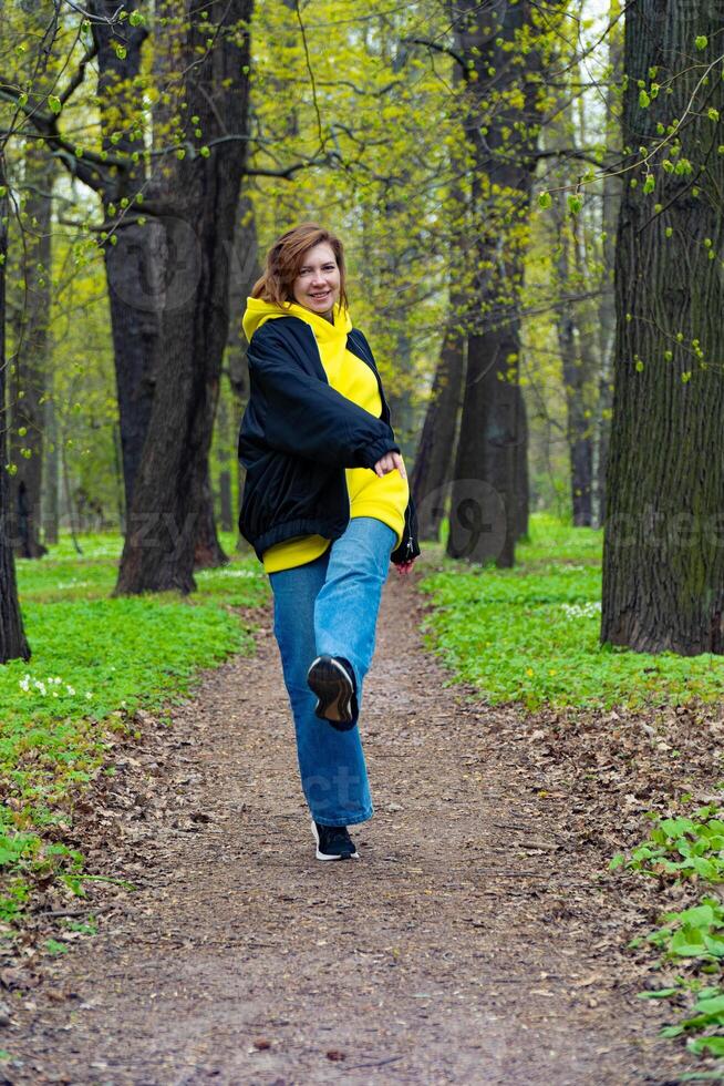 A young woman in a yellow hoodie and blue jeans enjoys a leisurely walk in a lush green park during spring, surrounded by budding trees photo