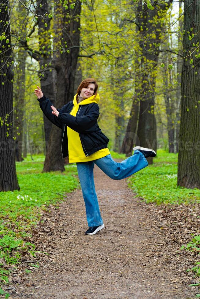 A young woman in a yellow hoodie and blue jeans enjoys a leisurely walk in a lush green park during spring, surrounded by budding trees photo