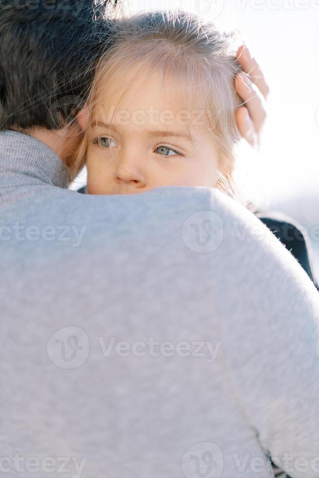 Little girl peeks out from behind her daddy shoulder while sitting in his arms. Back view photo