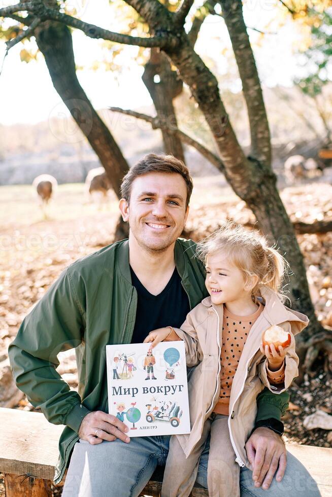 sonriente papá con un pequeño niña en su regazo se sienta en un banco con un libro en su manos. subtítulo. mundo alrededor foto